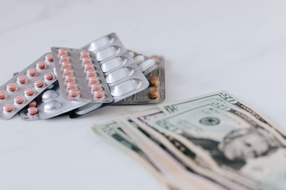 Photograph of several blister packs of pink and white pills arranged on a white surface, with a few loose pills scattered around. In front of the medication, there are multiple US dollar bills, partia