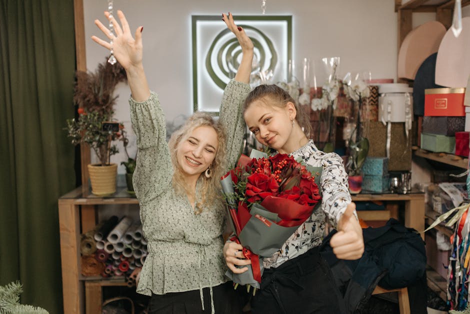 Two young women in a florist shop celebrating with a floral gift. One woman, with curly blonde hair and wearing a green patterned blouse, is smiling broadly with her arms raised in excitement. The oth