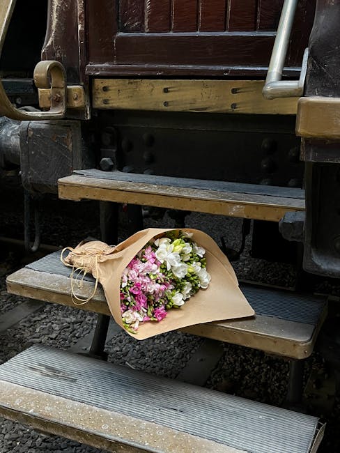 A vibrant bouquet of fresh flowers resting on a wooden train carriage step. The arrangement features white lilies, pink carnations, and small pink blooms, wrapped in beige craft paper with a rustic tw