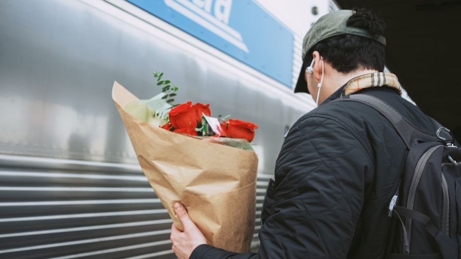 A person wearing a grey cap, black jacket, white earphones, and carrying a black backpack is holding a bouquet of fresh red roses wrapped in brown kraft paper. The bouquet features vibrant red roses w
