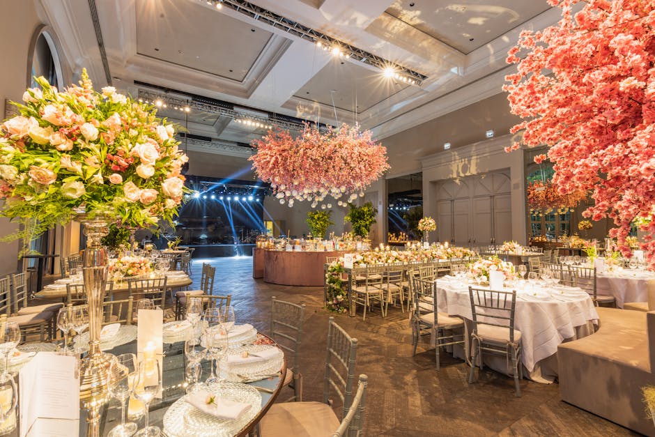 An elegantly decorated wedding reception hall featuring floral arrangements and decor by flowersplaistow.co.uk. The room showcases multiple round tables draped with white tablecloths, each adorned wit