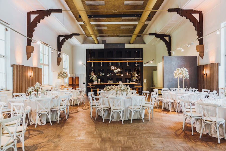 An elegantly decorated event space featuring round tables draped with white tablecloths, each adorned with floral centerpieces composed of white and blush pink roses, peonies, and greenery arranged in