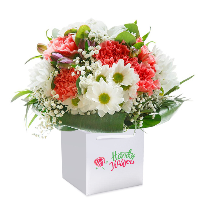 This striking bouquet, brought to life by a skilled local florist in Plaistow, is a harmonious blend of energy, softness, and connection to the area. The central display features a medley of ruffled coral and deep red carnations, their plush textures densely layered and evocative of festive gatherings in Plaistow's community hubs. Interspersed white daisy chrysanthemums with radiant lime-green centers provide bright, hopeful accents, while airy, cloud-like sprigs of baby's breath drift through the composition with a sense of grace. A discreet hint of purple within a few closed buds adds a layer of sophistication and fresh promise, reminiscent of the ever-unfolding changes in nearby Plaistow Park. Large, shiny green leaves form a natural frame, their hearty presence grounding the display and replicating the park's lush surrounds. The rounded arrangement stands firmly in a pure white gift bag, completed by elegant yet sturdy ribbon handles-a nod to convenience and modern style. Captured against a seamless white background, every nuance of colour, texture, and craftsmanship is in focus, underlining the bouquet's vitality and premium preparation. Whether chosen for a special Plaistow birthday, anniversary, or a spontaneous act of kindness, this bouquet promises to deliver heartfelt joy straight from the heart of the neighbourhood.